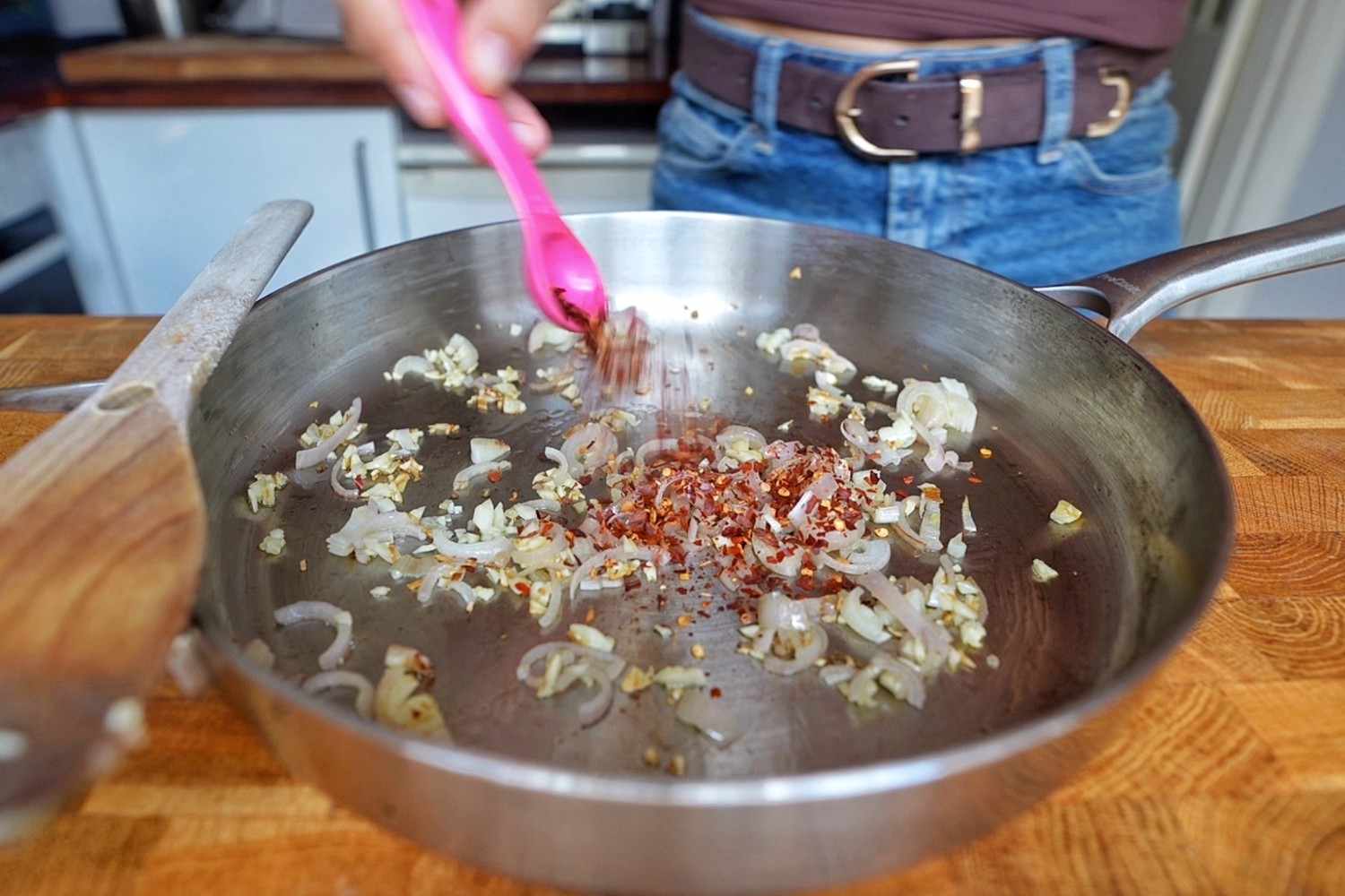 Emily adding red pepper flakes to softened sliced shallots and garlic in a stainless steel skillet.