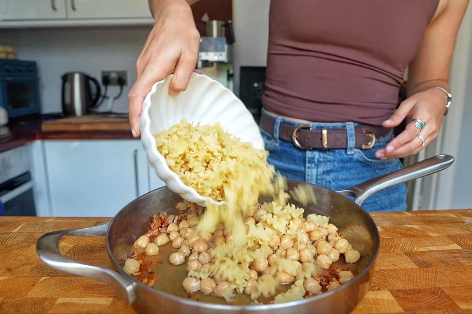 Pouring dry mafaldine pasta from a white scalloped bowl into a skillet of chickpeas.