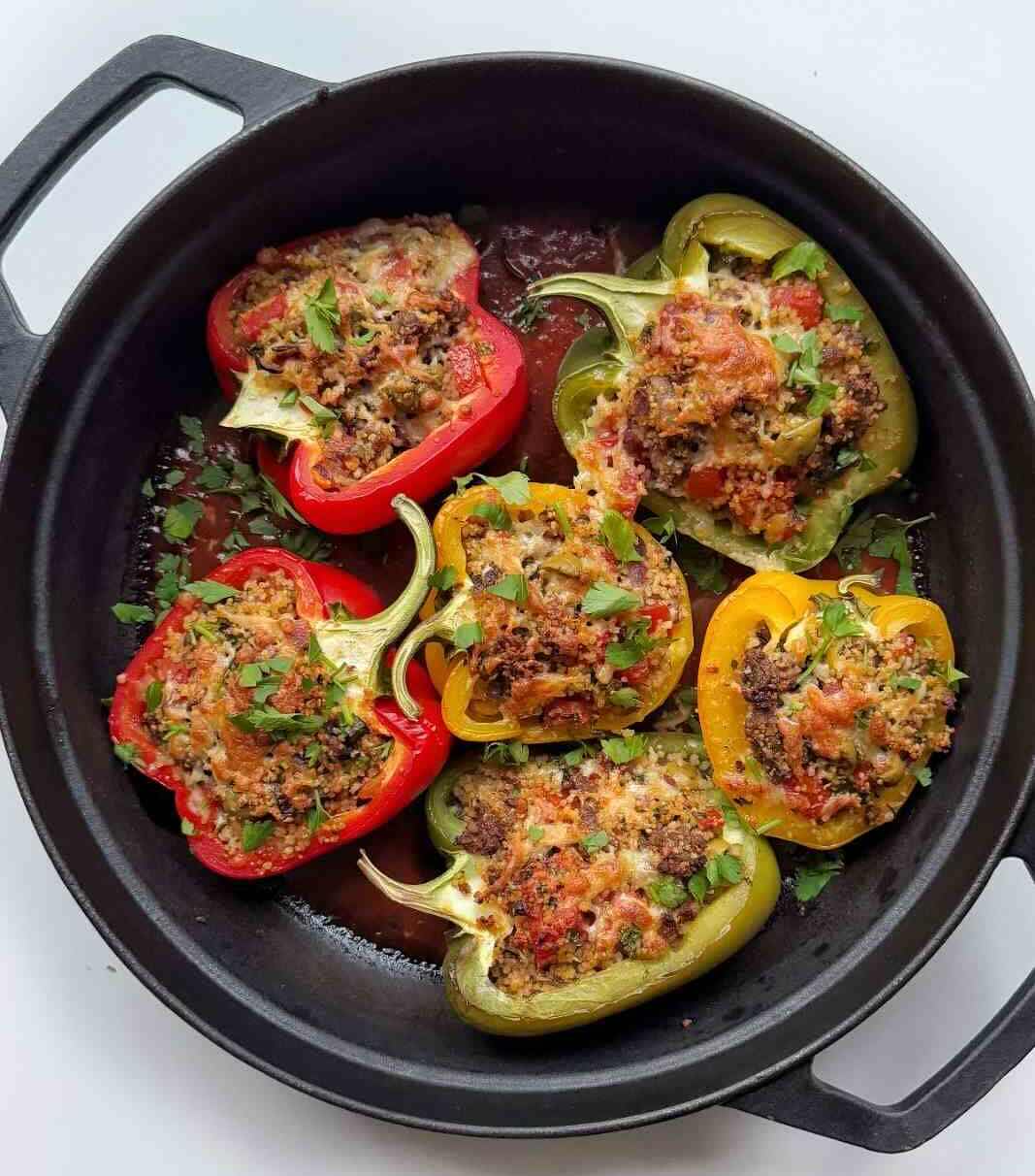 Six ground turkey stuffed peppers in red, yellow and green, filled with couscous and topped with melted cheese and parsley, in a cast iron pan.
