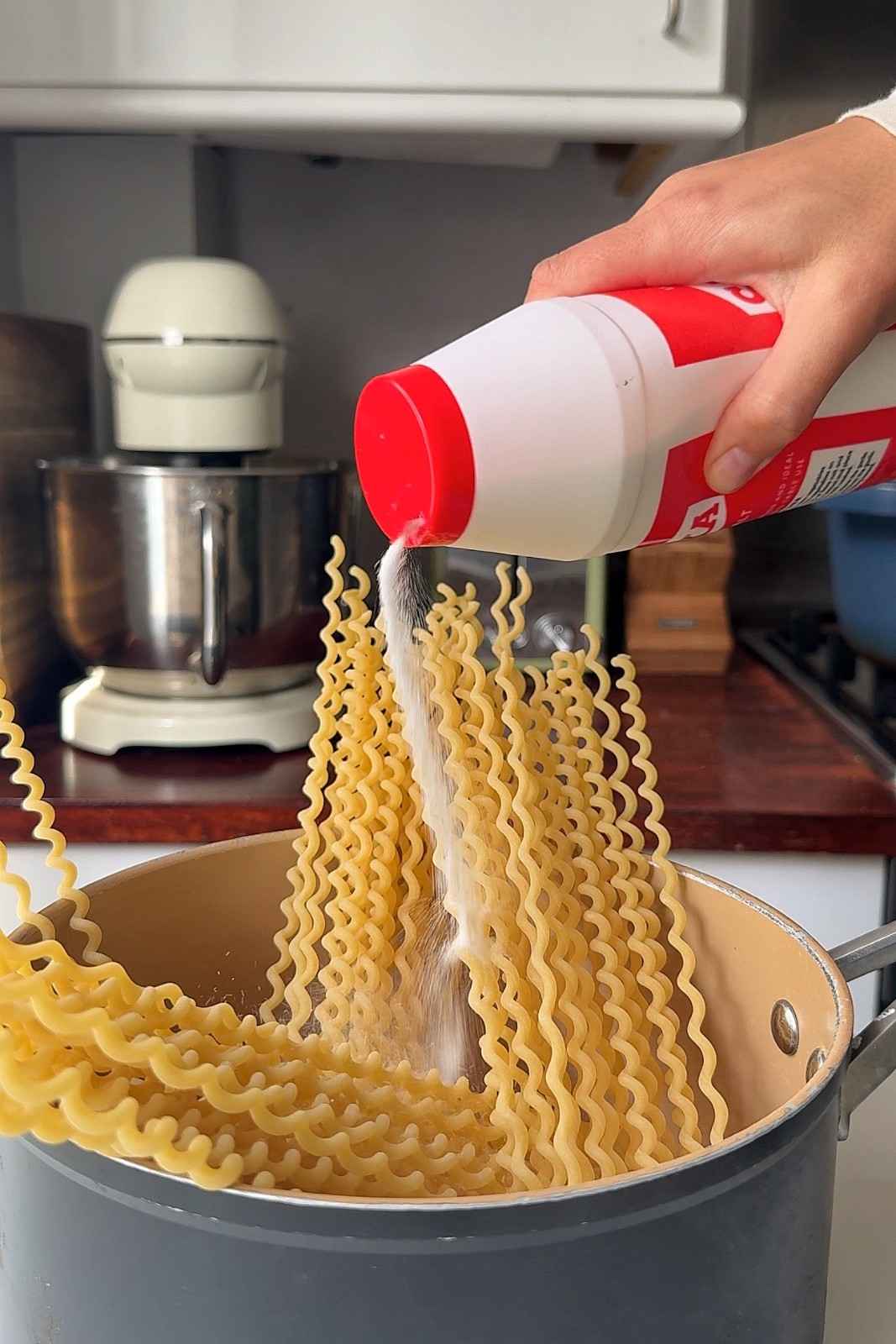Salt being poured into the pot as long wavy pasta goes in to cook.