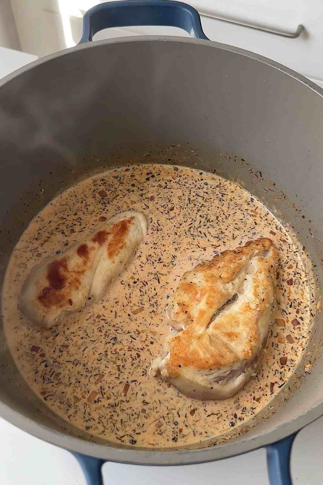 Two seared chicken breasts simmering in a creamy peppercorn sauce flecked with herbs and whole peppercorns.