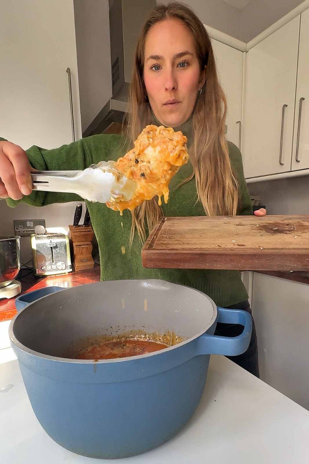 Emily lifting a sauce-coated chicken breast out of the pot with tongs onto a wooden cutting board.