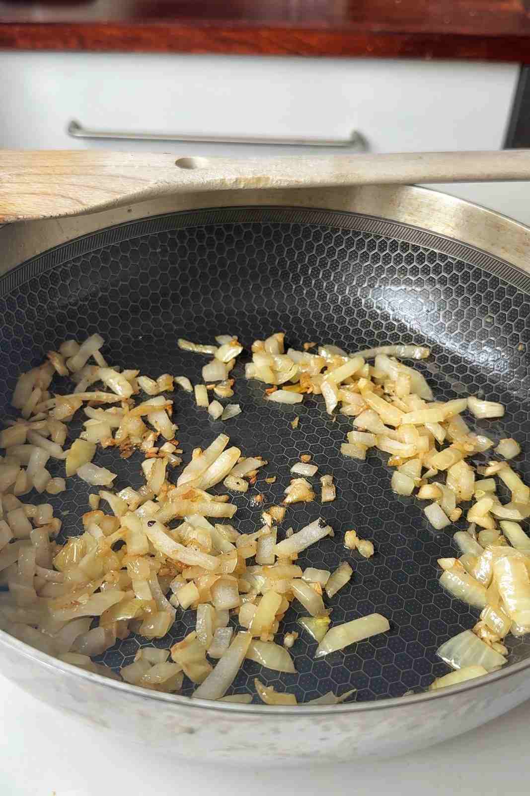 Diced onion and chopped garlic softened and lightly golden in a deep skillet, with a wooden spatula resting across the pan.