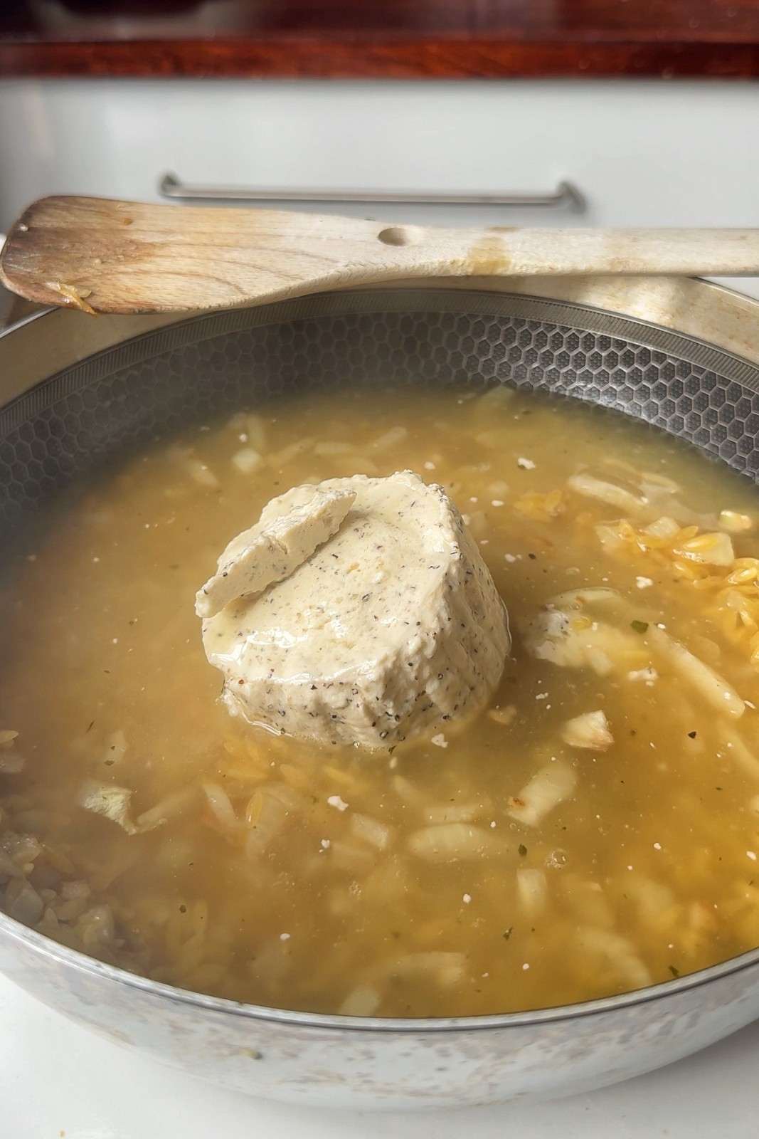 A whole Boursin cheese sitting in chicken broth with orzo and onion, ready to be broken down into the sauce.