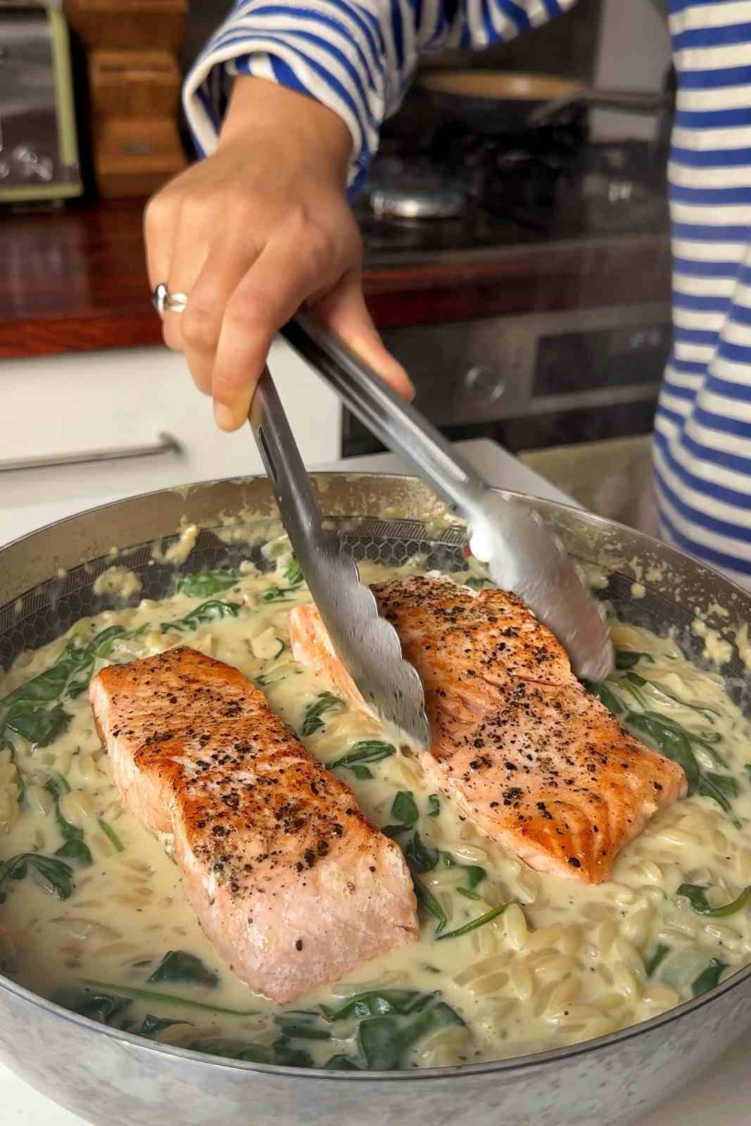 Tongs placing seared salmon fillets on top of creamy orzo and wilted spinach in the skillet.