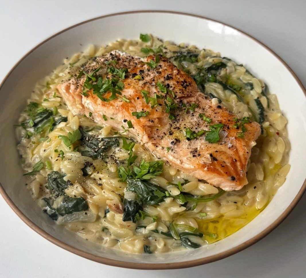 A bowl of one pan creamy Boursin salmon orzo with wilted spinach, topped with a seared salmon fillet, chopped parsley and cracked black pepper.