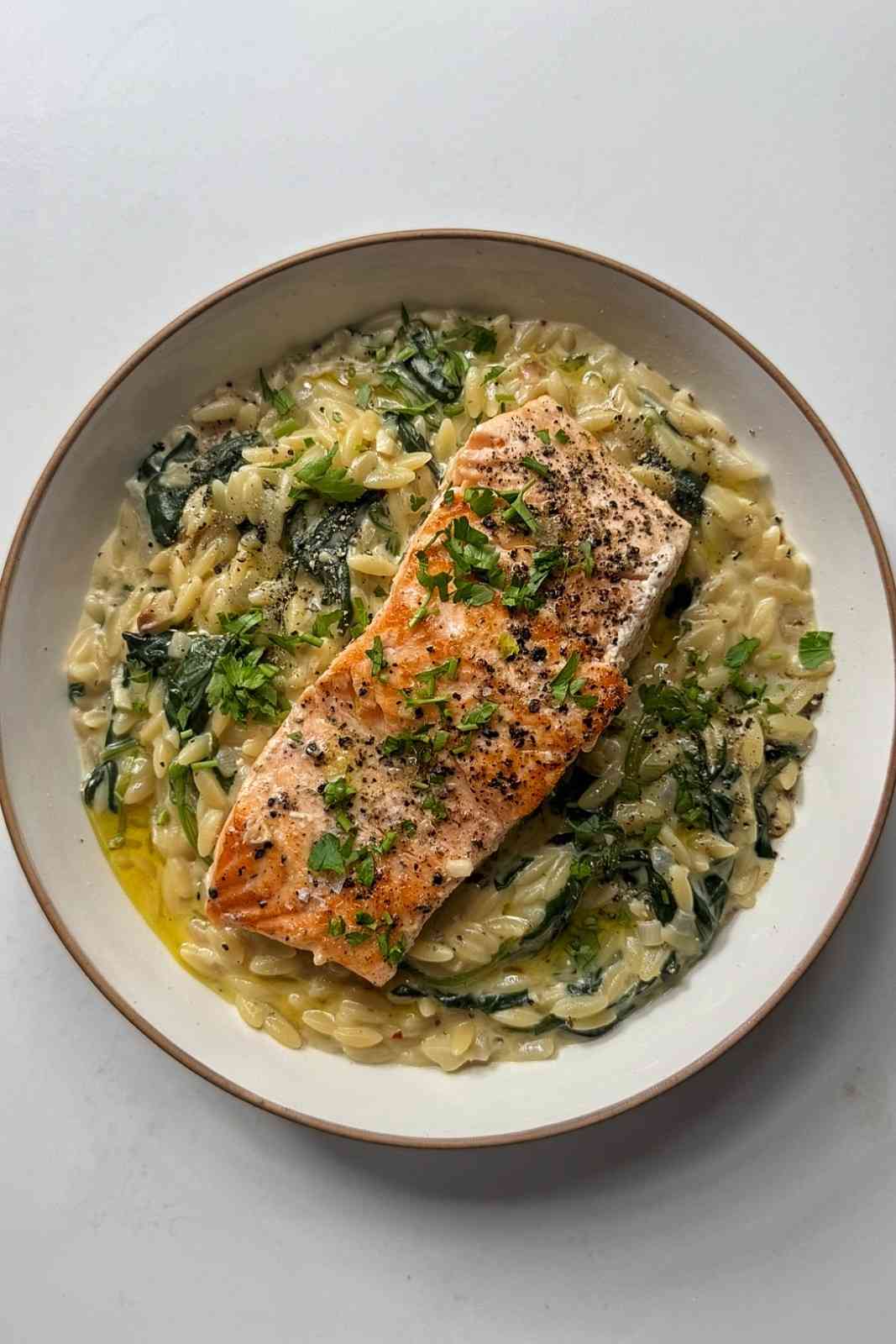 Overhead view of creamy Boursin salmon orzo in a white bowl, with a seared salmon fillet, wilted spinach and fresh parsley.