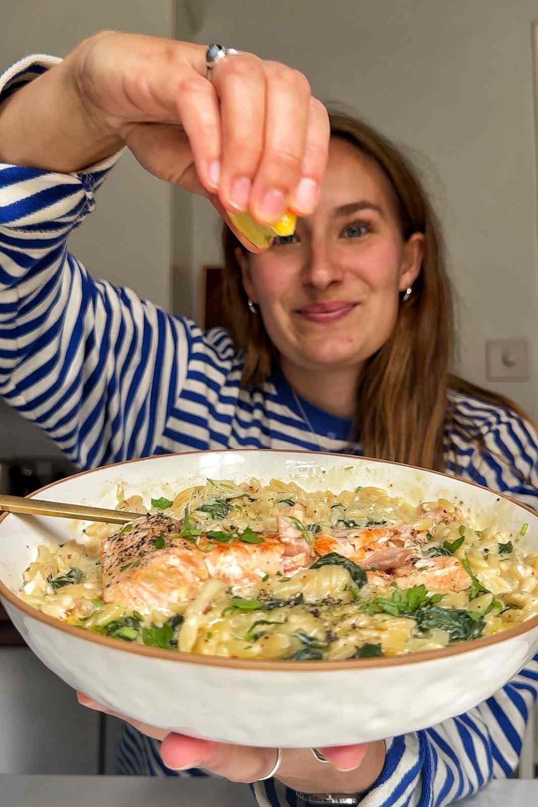 Emily Roz squeezing a lemon wedge over a bowl of creamy Boursin salmon orzo.
