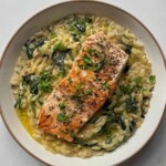 Overhead view of creamy Boursin salmon orzo in a white bowl, with a seared salmon fillet, wilted spinach and fresh parsley.