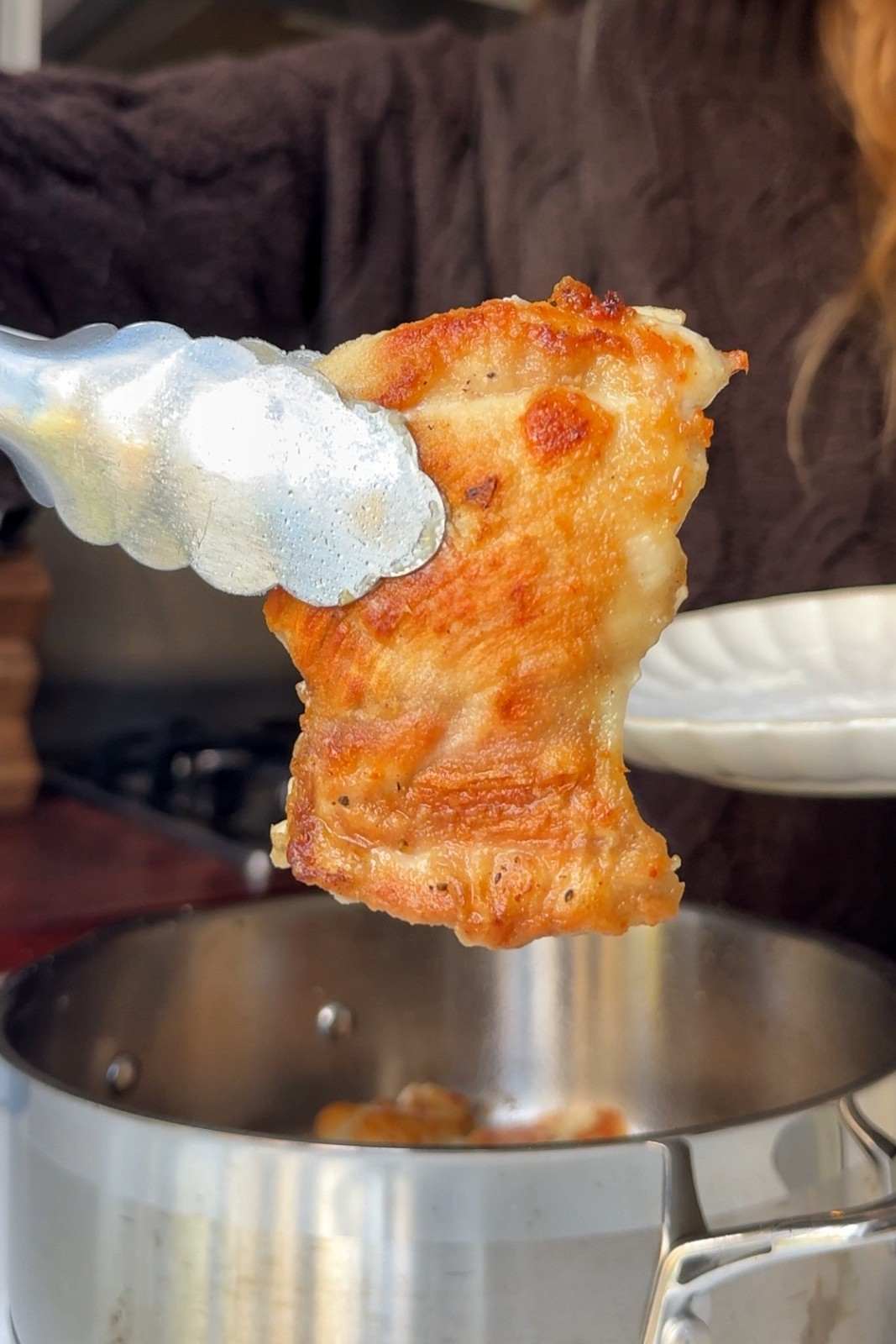Tongs lifting a golden, flour-crusted chicken thigh out of a stainless steel pan after searing.