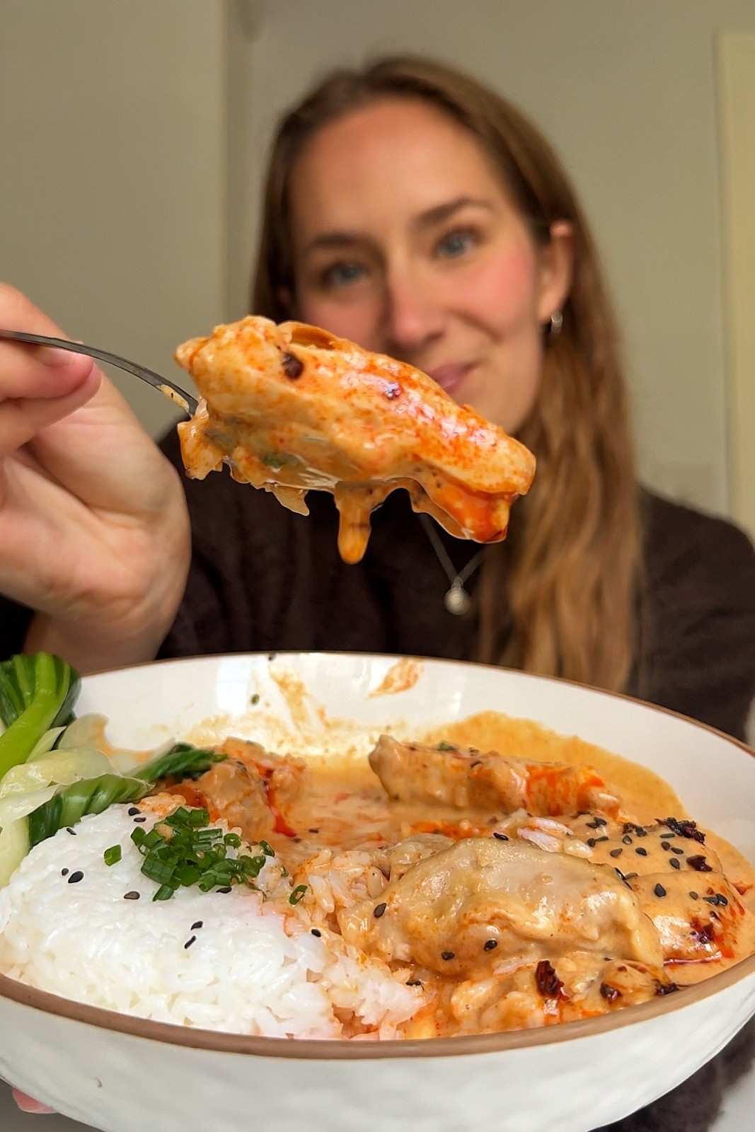 Emily holding a forkful of honey sriracha chicken up to the camera over a bowl of jasmine rice, bok choy and creamy orange sauce.