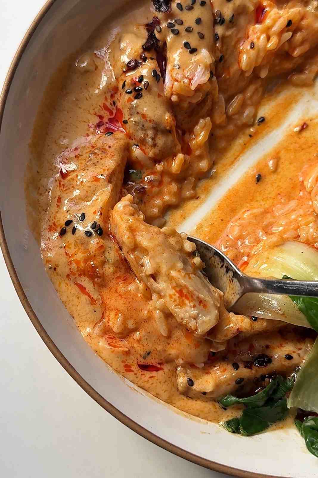 Close-up of a bowl of honey sriracha chicken with jasmine rice and bok choy in creamy coconut sauce, topped with black sesame seeds, with a spoon resting in the bowl.