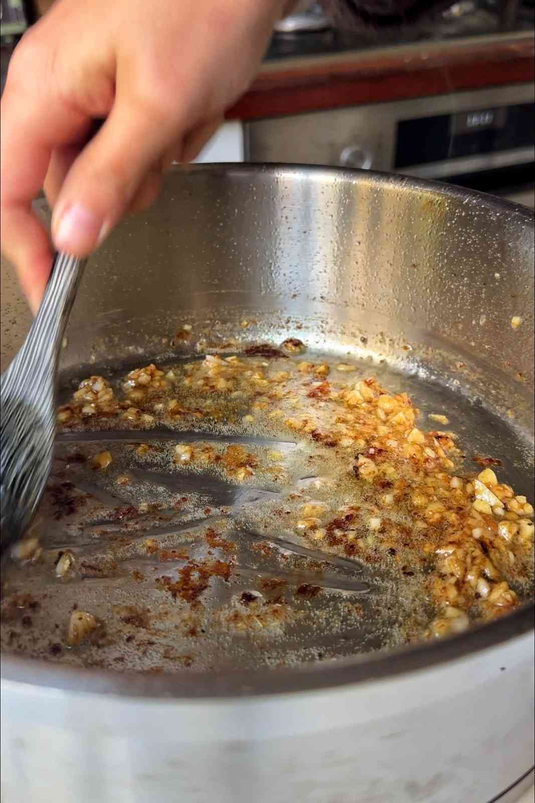 Minced garlic sizzling in melted butter in a stainless steel pan, with the browned bits from the chicken being scraped up with a spatula.