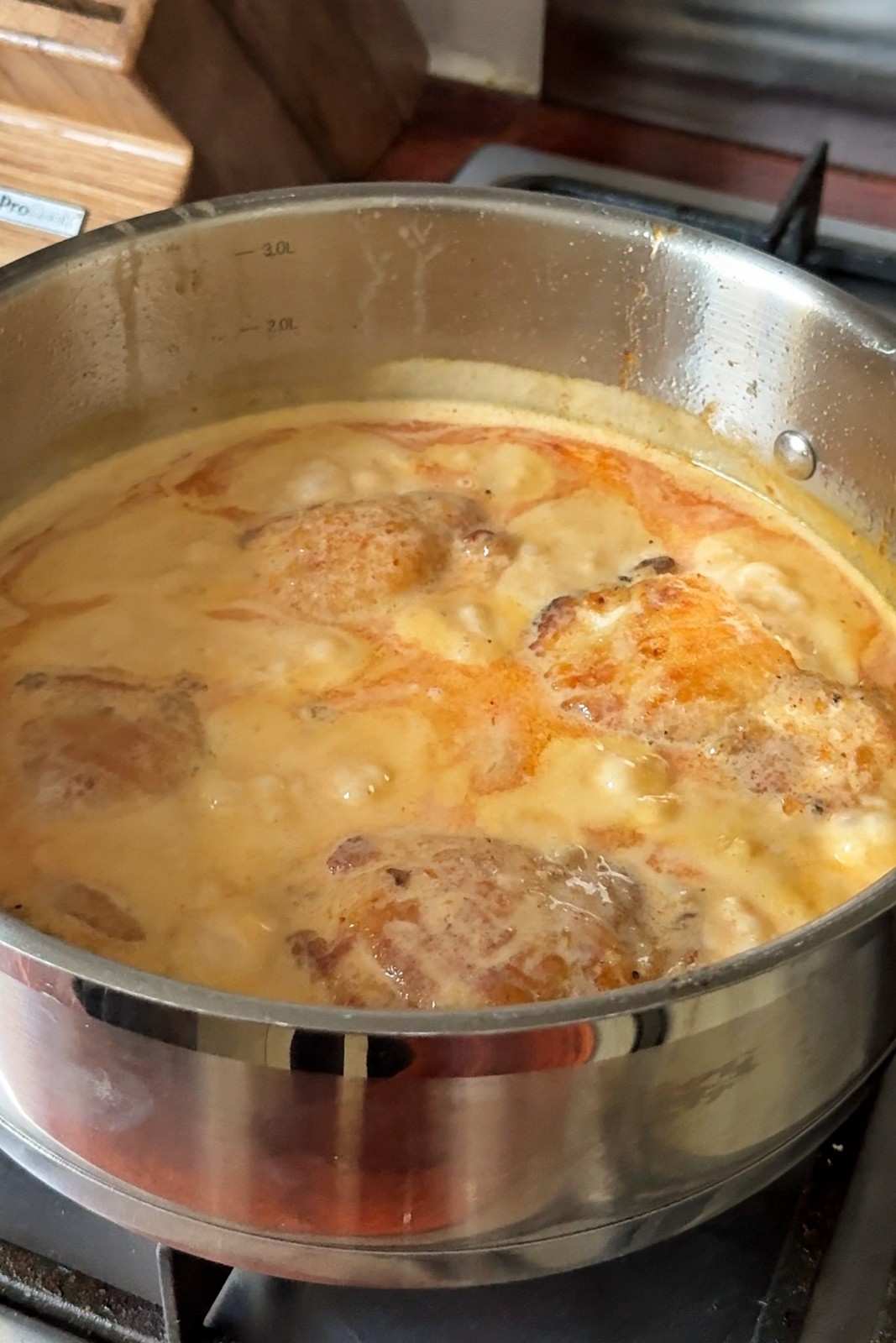 Chicken thighs bubbling in honey sriracha coconut sauce in a deep stainless steel pan on a gas stove.