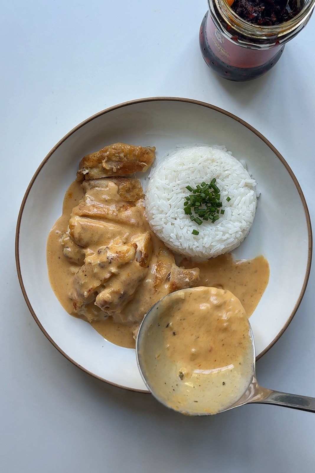 A ladle pouring creamy honey sriracha sauce over sliced chicken thighs beside a dome of jasmine rice topped with chives, with a jar of chili crisp in the background.