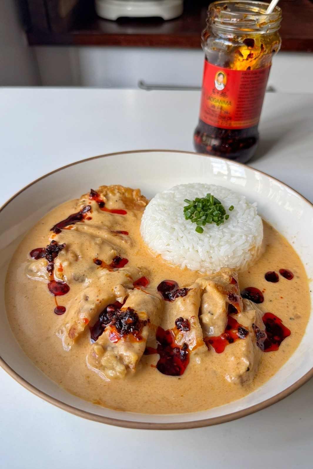 Creamy honey sriracha chicken on a white plate, sliced thighs drizzled with chili crisp next to a dome of jasmine rice topped with chives, with a jar of chili crisp behind.