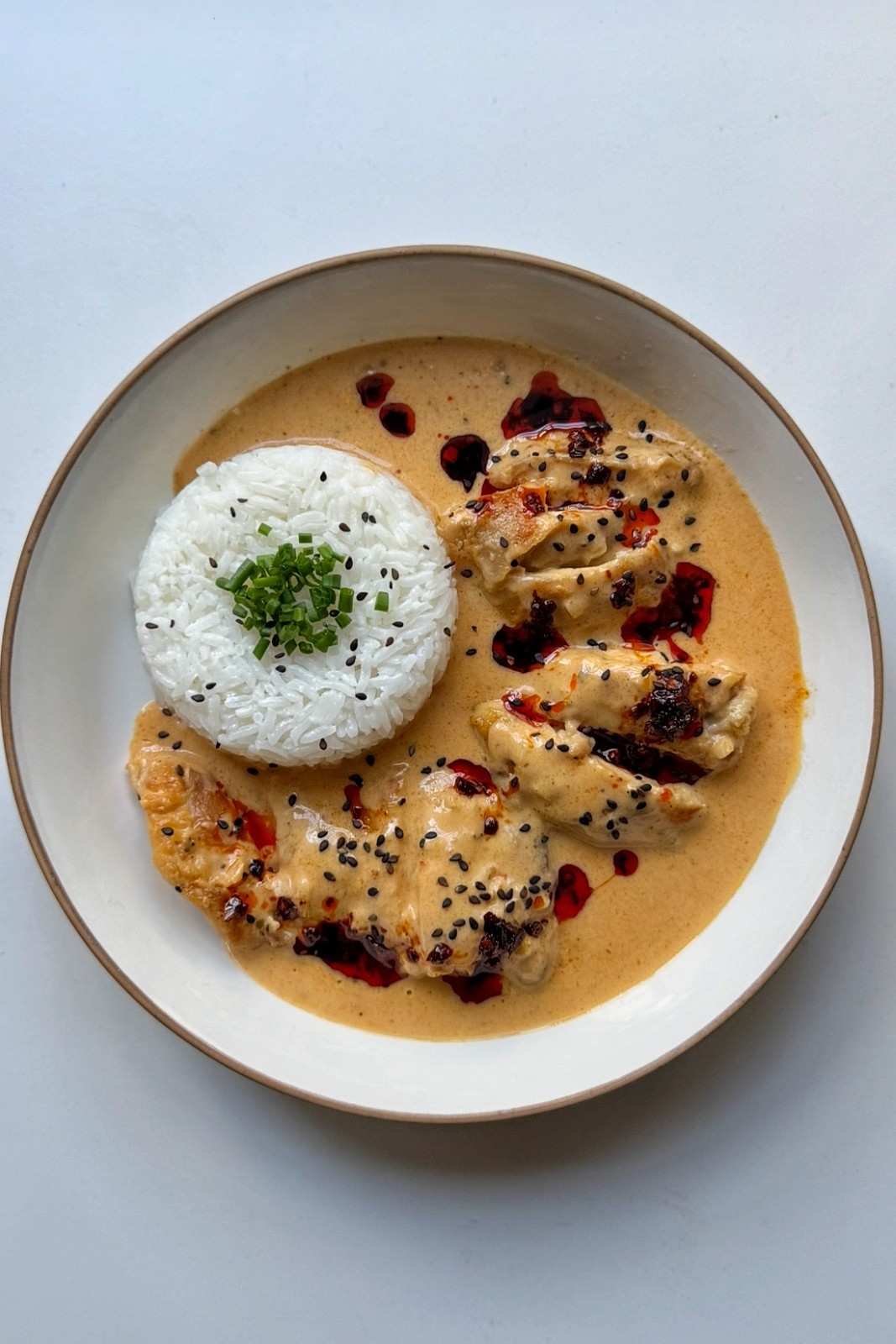 Overhead view of creamy honey sriracha chicken on a white plate, sliced chicken thighs in coconut sauce drizzled with chili oil and black sesame seeds, beside a dome of jasmine rice topped with chives.
