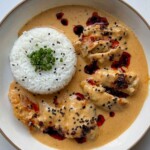 Overhead view of creamy honey sriracha chicken on a white plate, sliced chicken thighs in coconut sauce drizzled with chili oil and black sesame seeds, beside a dome of jasmine rice topped with chives.