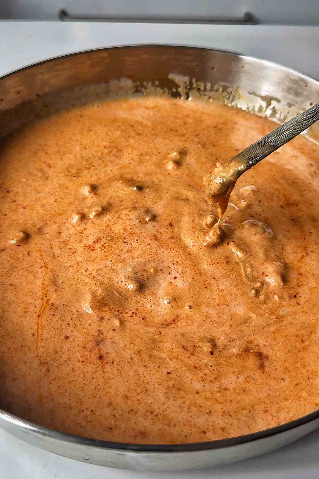 Creamy orange satay sauce simmering in a stainless steel skillet, with a spoon lifting some out to show its thickness.