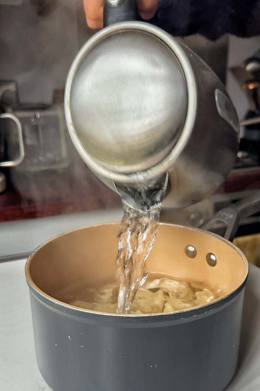 Boiling water being poured from a stainless steel kettle over gyoza in a dark nonstick saucepan.