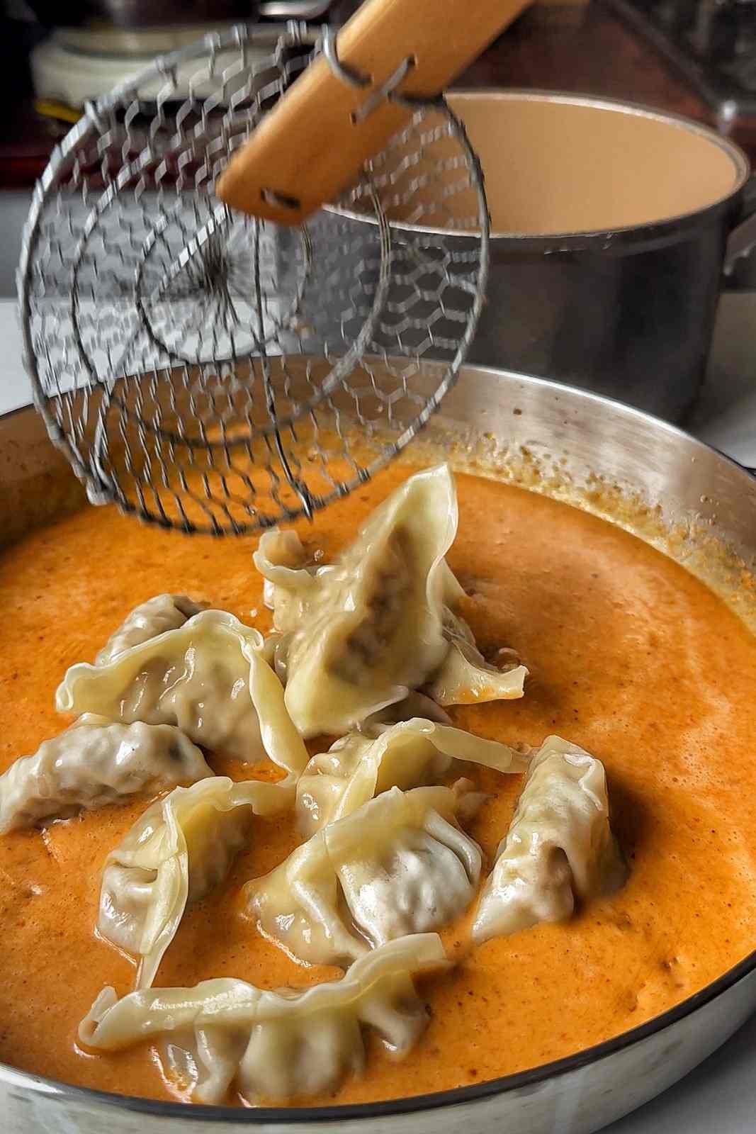 A wire spider strainer lowering boiled gyoza into a pan of creamy peanut satay sauce.