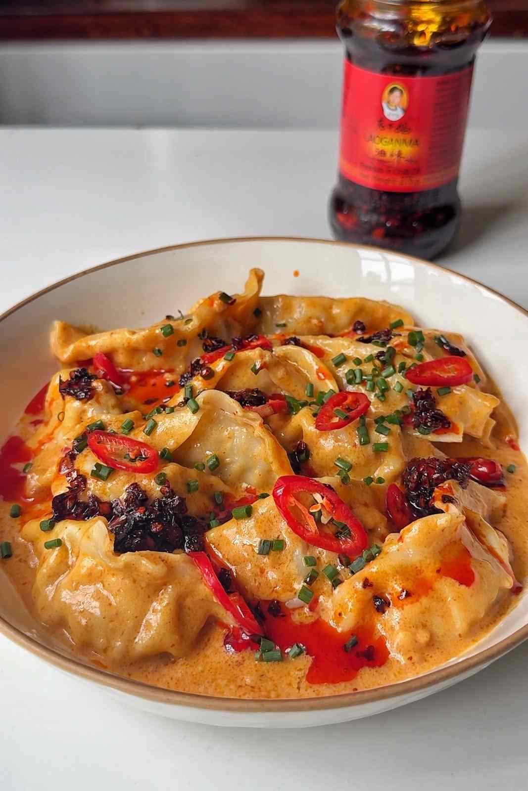 A white bowl of gyoza in creamy peanut satay sauce, topped with sliced red chilies, chopped chives and chili crisp, with a jar of chili crisp behind it.