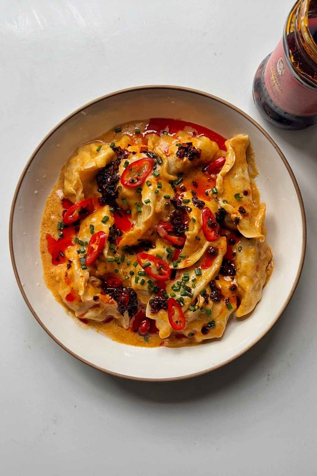 Overhead view of satay sauce dumplings in a white bowl, topped with sliced red chilies, chopped chives and chili crisp.
