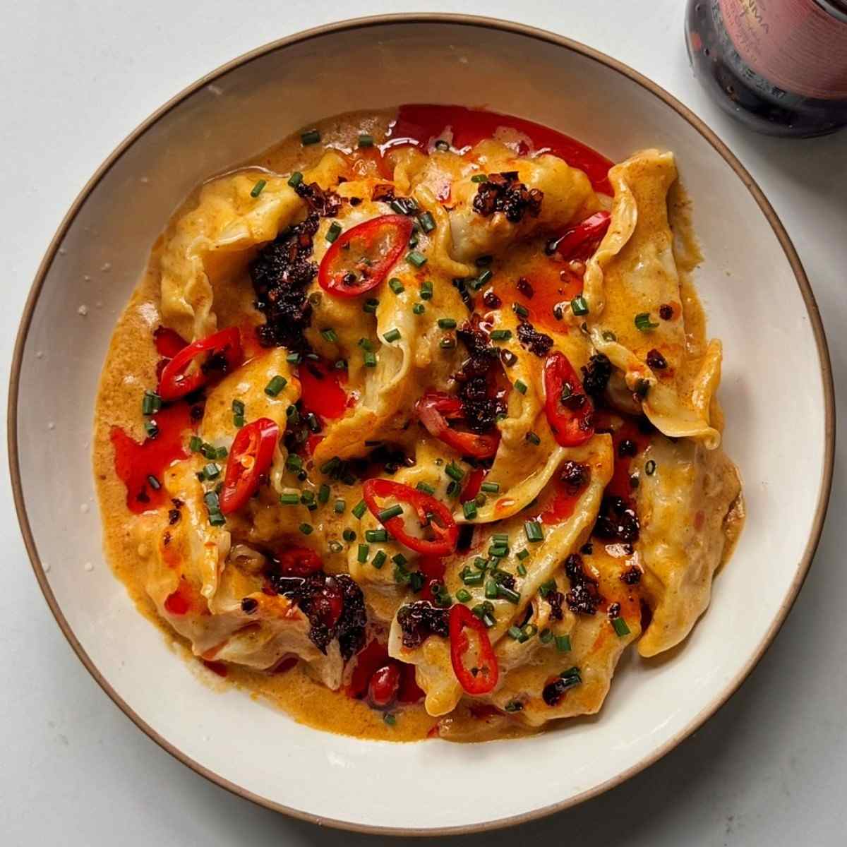 Overhead view of peanut satay sauce dumplings in a white bowl, topped with sliced red chilies, chopped chives and chili crisp.