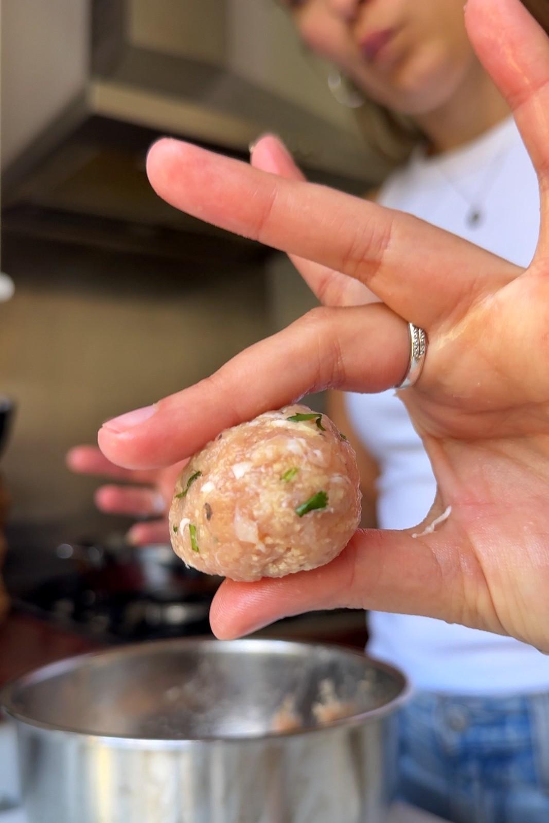 Emily Roz holding a raw chicken meatball flecked with chopped cilantro over the mixing bowl.