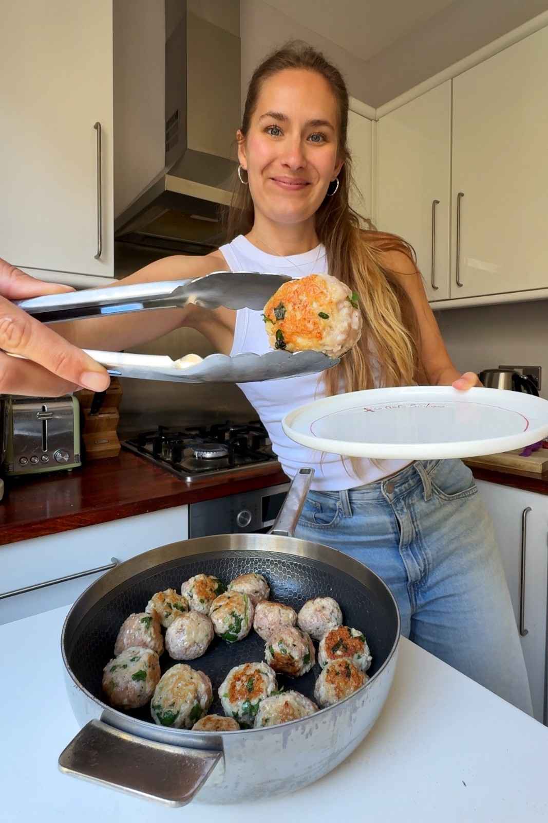 Emily holding up a golden seared chicken meatball with tongs over a pan of lightly browned meatballs, with a plate ready to set them aside.