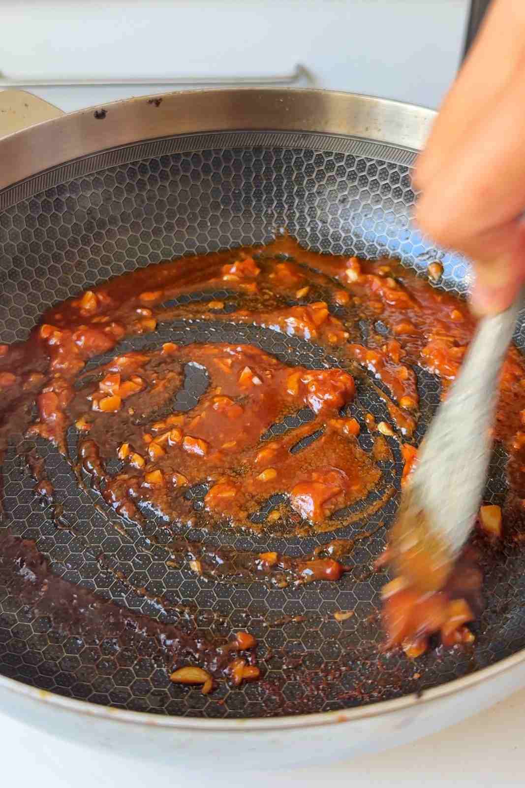 Garlic, miso paste, sriracha and soy sauce being stirred into a glossy orange paste in a nonstick pan.