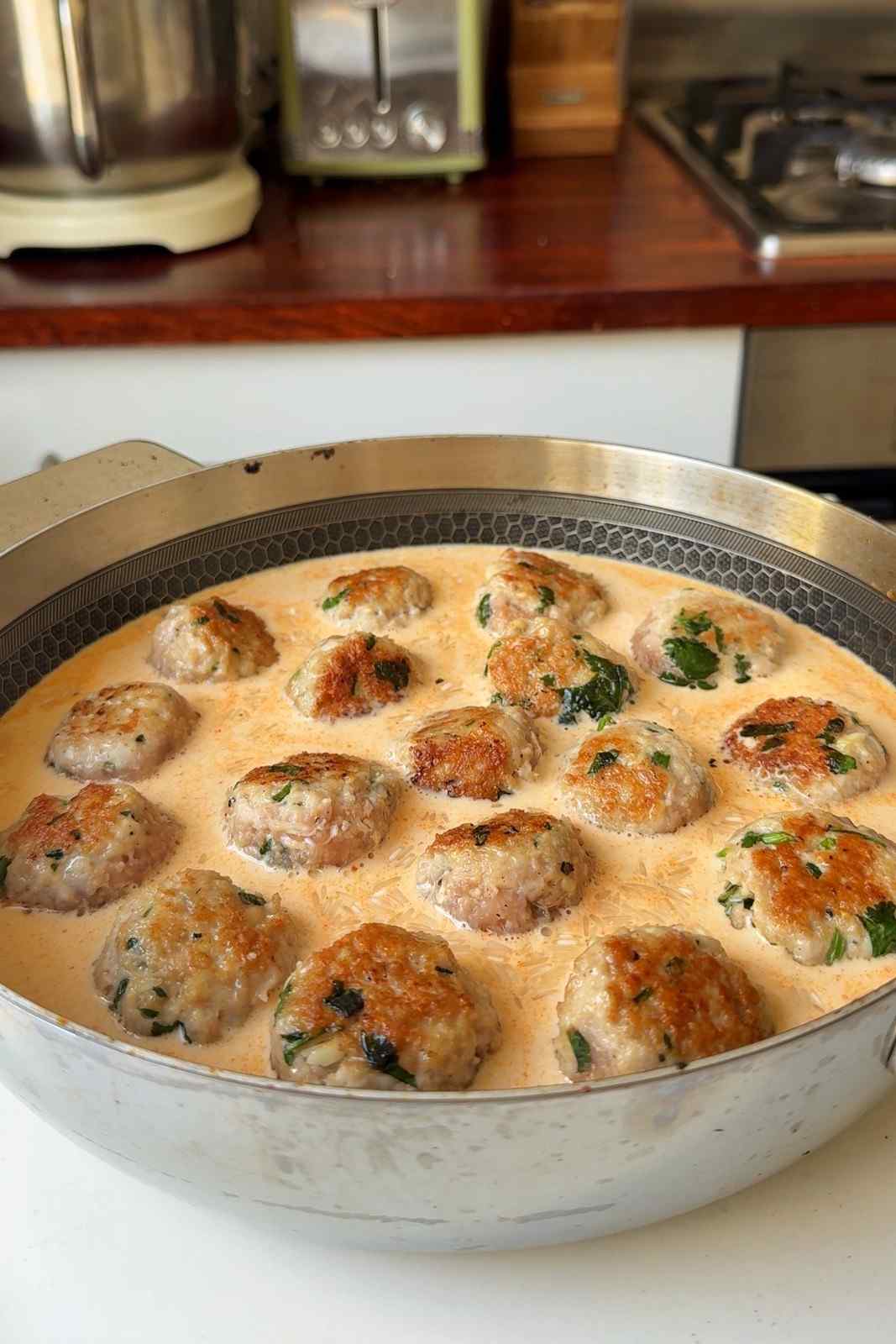 Seared chicken meatballs nestled into uncooked rice in a pale orange mix of cream and chicken stock, ready for the lid to go on.
