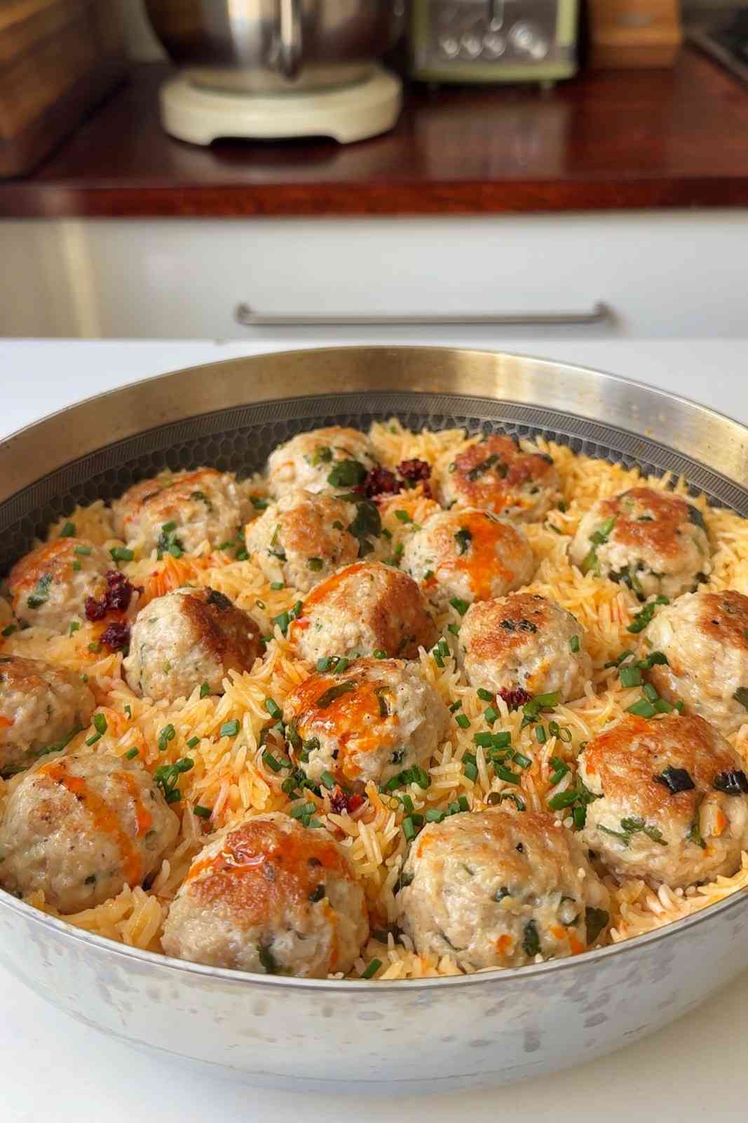 One pot spicy miso chicken meatballs and rice in a wide stainless steel pan, with golden seared meatballs sitting in fluffy orange-tinted rice and scattered with chopped chives.