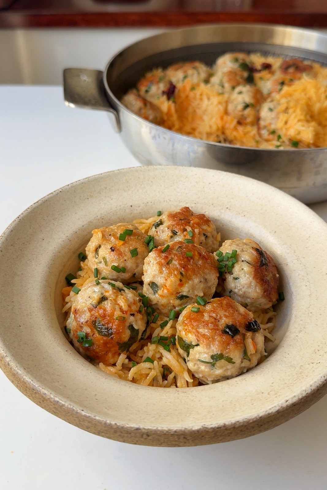 A bowl of chicken meatballs flecked with cilantro over creamy orange-tinted rice, topped with chopped chives, with the pan of rice behind it.