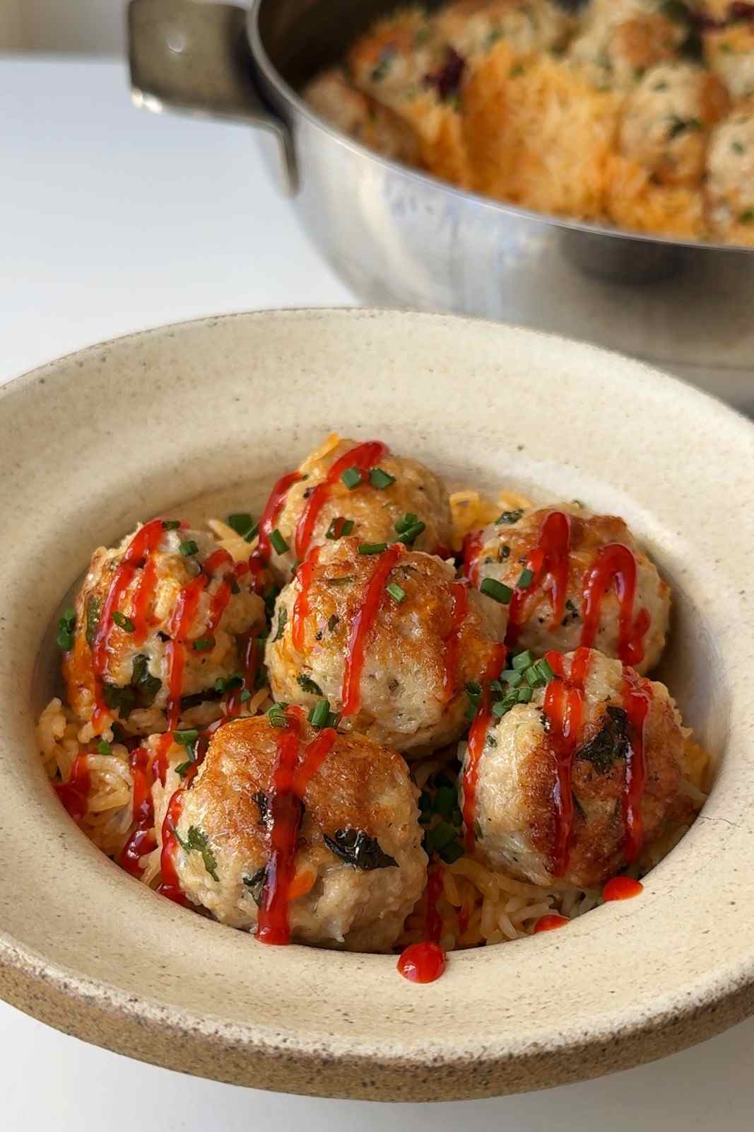 Spicy miso chicken meatballs on creamy orange-tinted rice in a speckled stoneware bowl, drizzled with sriracha and topped with chopped chives, with the pan of meatballs and rice behind.