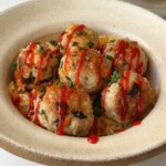 Spicy miso chicken meatballs on creamy orange-tinted rice in a speckled stoneware bowl, drizzled with sriracha and topped with chopped chives, with the pan of meatballs and rice behind.