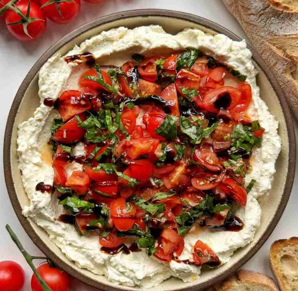 A plate of bruschetta dip recipe made with whipped cream cheese and feta topped with garlicky basil tomatoes with olive oil and a balsamic glaze.